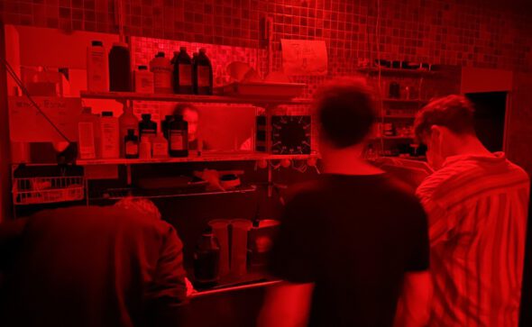 people in a red-lit photography lab, standing in front of a wet bench with chemicals